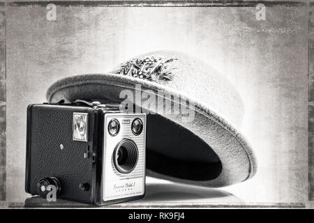 Black & white close up of old 1950's Fedora/ trilby hat leaning on vintage Kodak Box Brownie camera (Model 1) of same period. Aged effect background. Stock Photo