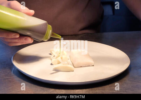 Close up of male chefs hands pouring green sauce from a clear plastic bottle onto a white plate with white fish on it, the plate is on a silver metal  Stock Photo