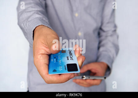 Cropped short of young man reaching out hand with blue credit card with fingerprint sensor and holding mobile in other hand. Businessman giving paymen Stock Photo