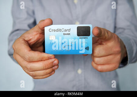Young man stands at white background and holds credit card with fingerprint sensor in one hand and points with index finger of other hand to it. Purch Stock Photo