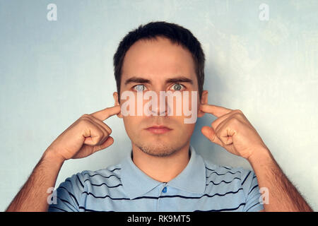 Handsome young male model plugs ears with displeased expression as hears annoying loud noise or roaring not far from him, over blue concrete wall. Not Stock Photo