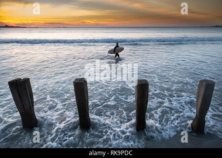 surfer leaves the water at sunset. Shot from the shoreline with groynes in the foreground Stock Photo