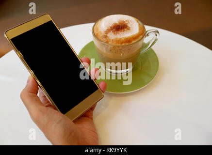 Man holding cellphone in his hand beside a cup of hot cappuccino coffee on the white table Stock Photo