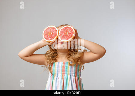 Cheerful little girl child is having fun and substitutes halves of grapefruit instead of eyes. Gray background, studio Stock Photo