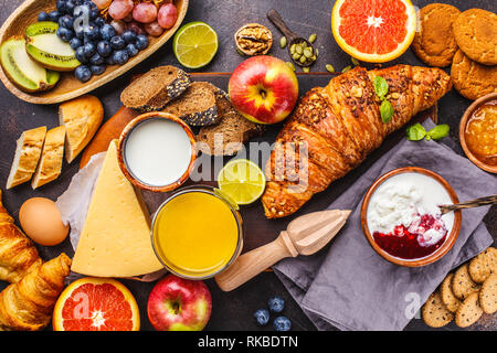 Healthy balanced breakfast on a dark background. Muesli, milk, juice, croissants, cheese, biscuits and fruit, top view. Stock Photo