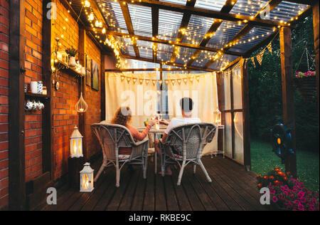 Young couple man and woman sit on white rattan garden chairs on wooden romantic outdoor terrace in the evening celebrating and toasting, spending time Stock Photo