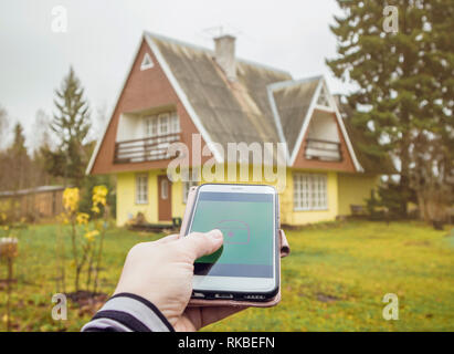 Person hand press lock/unlock button from distance on smartphone app to open/close rural home house door. Modern lifestyle technology concept. Stock Photo