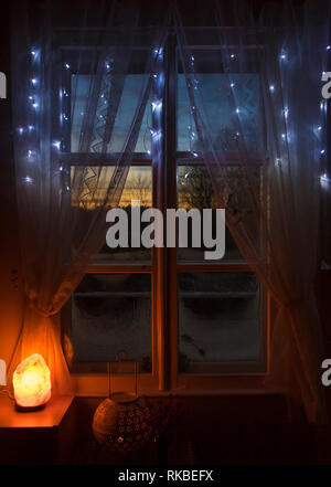 Decorative Himalayan Salt lamp turned on by old white wooden window with white net fabric curtains, looking out the wooden frames window sun setting o Stock Photo