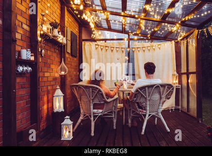 Young couple man and woman sit on white rattan garden chairs on wooden romantic outdoor terrace in the evening celebrating and toasting, spending time Stock Photo