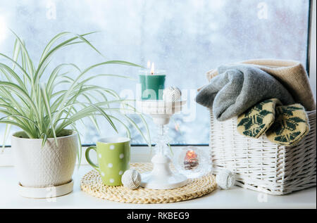 Cozy winter set on window sill. Romantic style tall candle holder, rattan basket with knitted mittens and warm plaids and blanket, snow falling outdoo Stock Photo