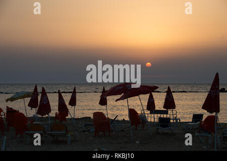 Warm summer sunset view of an urban dirty beach, full of garbage on the sand, chairs and umbrellas, some open, some closed, in red, seascape. Stock Photo