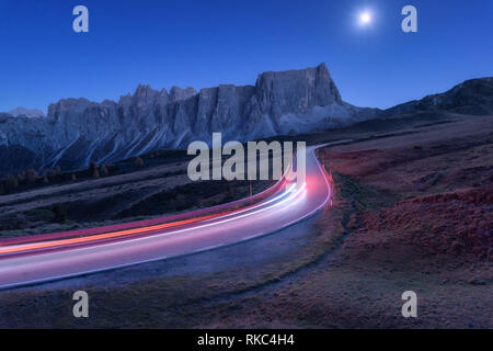 Blurred car headlights on winding road at night in autumn. Landscape with asphalt road, light trails, mountains, hills, blue sky with moonlight at dus Stock Photo