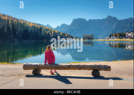 Young woman sitting on the wooden bench on the coast of Misurina lake at sunset in autumn. Dolomites, Italy. Landscape with girl in red jacket, reflec Stock Photo