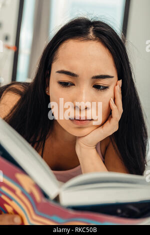 Dark-eyed Asian woman feeling involved in reading book Stock Photo