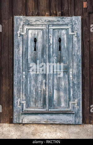 Traditional wood window shutters in blue flaking paint and artisan metal hinges set in borwn wood panel wall of alpine outbuilding Stock Photo