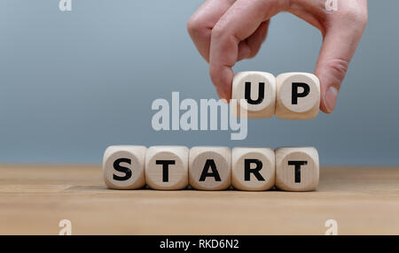 Cubes form the words 'START UP' while to fingers lift the letters 'UP' in the air. Cubes are on a wooden table in-front a grey background. Stock Photo