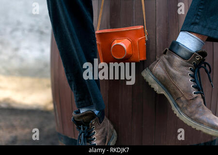 Man with leather boots sitting on wooden barrel and holding vintage film camera in a leather case Stock Photo