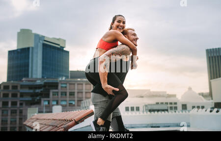 Side view of a fitness couple having fun while training on terrace of a building. Man carrying a fitness woman on his back walking on rooftop. Stock Photo