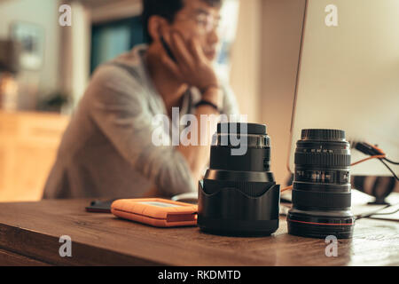 Two professional camera lenses and portable hard disk on table with photographer working on computer at the back. Photographer editing photos on deskt Stock Photo