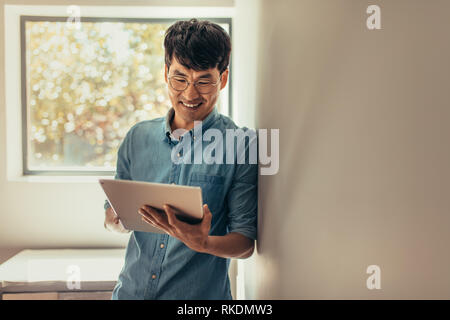 Smiling man leaning to a wall and using tablet pc. Man standing at home with digital tablet updating his social media status. Stock Photo