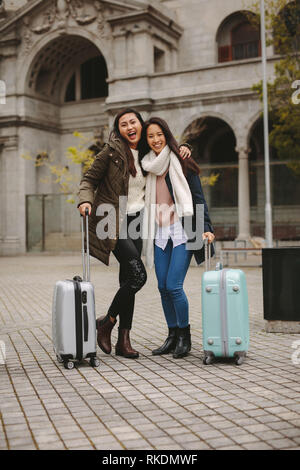 Two asian tourists exploring the city and having fun. Smiling female tourists standing together on street holding their luggage. Stock Photo