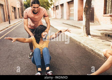 Excited young woman being pushing on skateboard by her boyfriend outdoors on street, with friends sitting by the street. Couple enjoying themselves ou Stock Photo