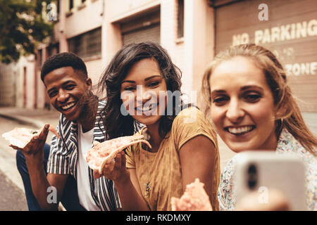 Woman taking selfie with friends while eating pizza. Group of men and women eating pizza sitting outdoors on city street and taking selfie. Stock Photo