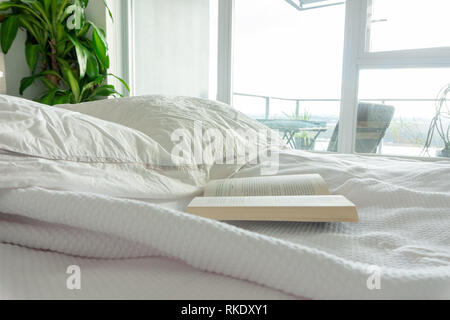 Reading in bed, staying in bed. Wrinkled sheets in a used bed, in a real home, vrbo or hotel bedroom. Apartment windows & balcony, with bright light. Stock Photo