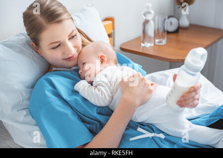 high angle view of smiling young mother holding baby bottle with milk and lying in bed with adorable baby Stock Photo
