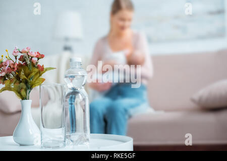 close-up view of glass, bottle of water, flowers in vase and mother breastfeeding baby behind at home Stock Photo