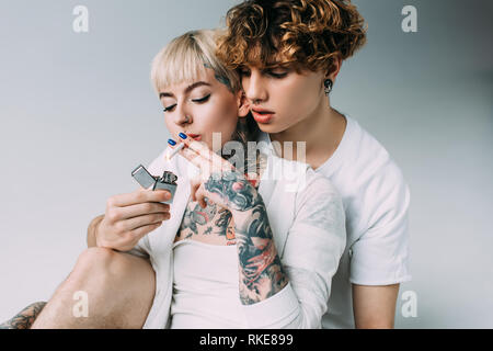 blonde tattooed girl smoking cigarette near handsome man with lighter in hand isolated on grey Stock Photo