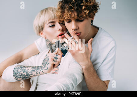 blonde tattooed girl holding lighter near man with cigarette in mouth isolated on grey Stock Photo
