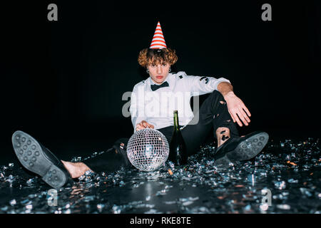 man with curly hair sitting in party cap with disco ball isolated on black Stock Photo