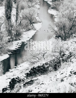 A snow-covered landscape, half frozen river with trees on the banks, flows through a shallow valley. Black and white photography Stock Photo
