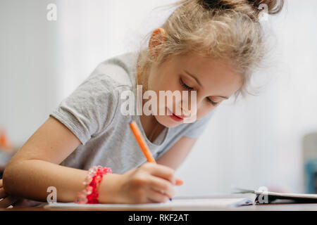 A seven-year-old girl sits at home at a table and writes in a notebook, completing a learning task or repeating lessons. Stock Photo