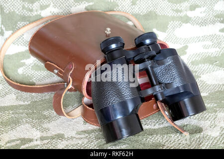 Vintage Porro prism black color military binoculars and brown leather carry case with strap on camouflage background side view close up Stock Photo