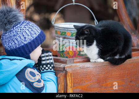 Little boy playing with a black cat. Black and white cat sitting on wooden well outdoors in the back yard Stock Photo