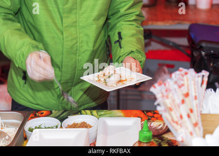 Chef makes portion of the food for his customer, paper coaster with dumplings and vegetable decoration in one hand, spoon in the other hand. Asian str Stock Photo
