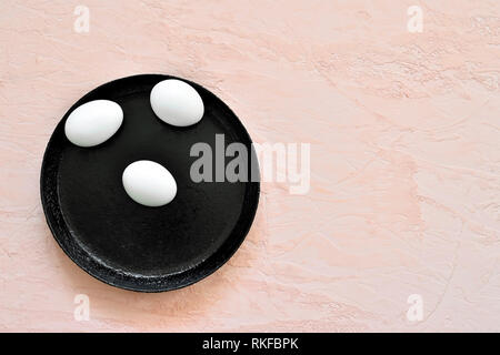 Three white eggs in a shell lie on a black pan against the background of a shade of living coral. Stock Photo