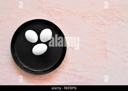 Three white eggs in a shell lie on a black pan against the background of a shade of living coral. Stock Photo