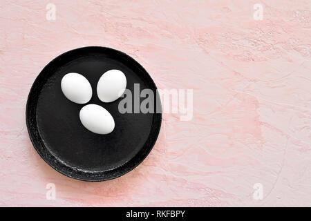 Three white eggs in a shell lie on a black pan against the background of a shade of living coral. Stock Photo
