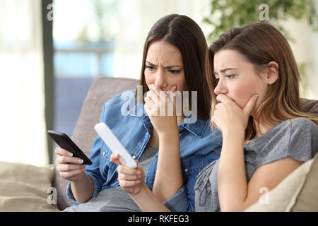 Two worried women checking smart phones content sitting on a couch in the living room at home Stock Photo