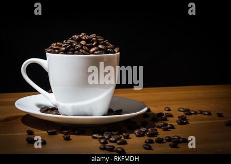 White cup filled with coffee beans  and coffee beans scattered on a wooden surface and on the white saucer. Stock Photo