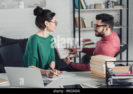 handsome man pointing with finger at book near attractive coworker in modern office Stock Photo