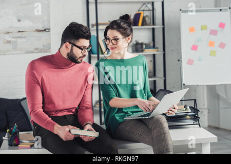 cheerful woman pointing with finger at laptop and looking at happy bearded man holding book Stock Photo