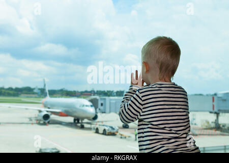 Little baby boy waiting boarding to flight in airport transit hall and looking through the window at airplane near departure gate. Active family Stock Photo