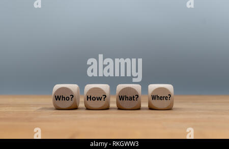Four cubes with the questions "who?", "how?", "what?" and "where?" are lying on a wooden desk in front of a grey background. Stock Photo
