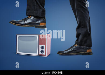 A giant businessman's leg stepping on a small retro TV box with an empty screen. Stock Photo