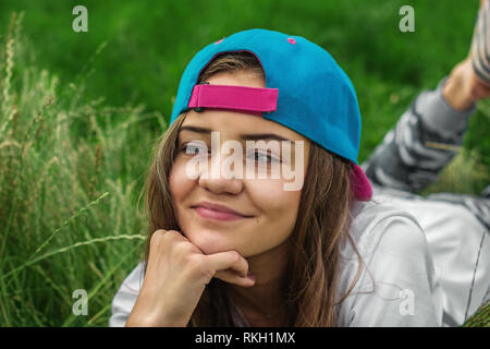 A beautiful teen girl in a baseball cap with her hair down lying on the grass looks away and smiles. Beginning of summer holidays for schoolchildren Stock Photo