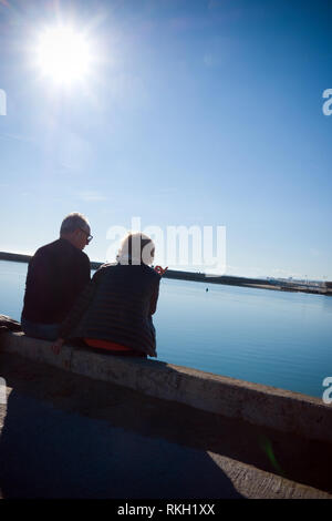 rear shot of an elderly couple discussing sit on a sea wall overlooking a calm harbour on a sunny winter's day in Carnon, France Stock Photo